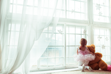 Balettiasuinen lapsi ison nallen kanssa katsoo ulos ikkunasta